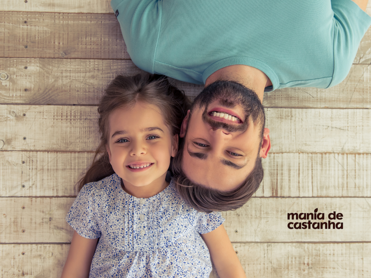 Pai deitado com a filha sorridente sobre piso de madeira clara – imagem para campanha de Dia dos Pais da Mania de Castanha